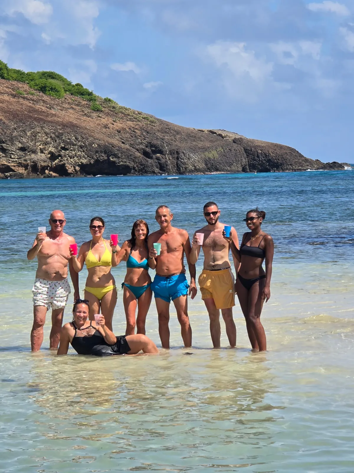 Groupe de vacanciers debout dans l'eau claire, gobelets levés, devant un îlet rocheux